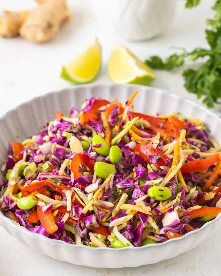 Asian cabbage salad with shredded red cabbage, broccoli and carrot mix, red bell pepper, edamame, roasted almonds, and fresh cilantro, tossed in a peanut-ginger dressing in a white bowl